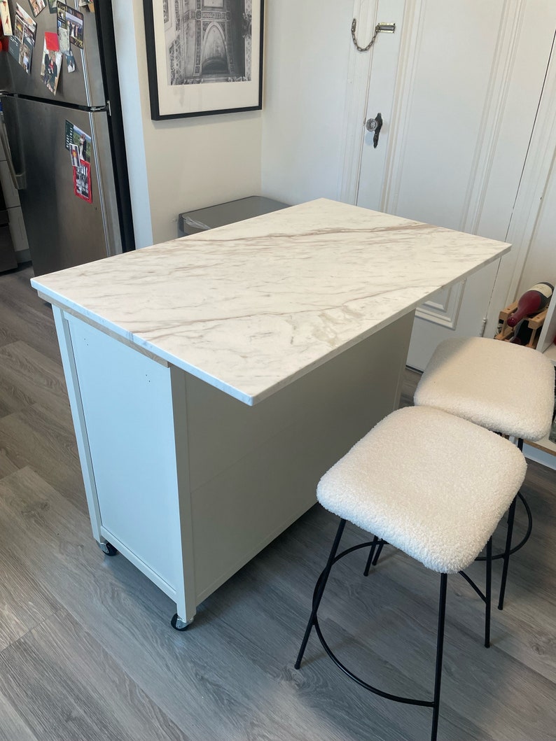 May include: White kitchen island with a marble countertop and two white upholstered bar stools with black metal legs.
