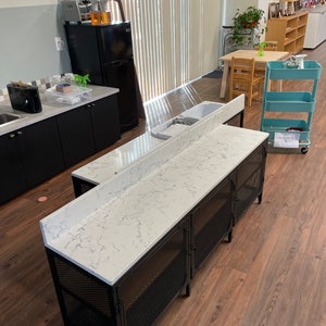 May include: A black metal industrial style cabinet with a white quartz countertop. The cabinet has three doors with wire mesh panels and is sitting on a wood floor.