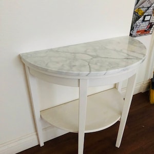 May include: White half-moon table with a marble top and a lower shelf. The table has four legs and is made of wood.