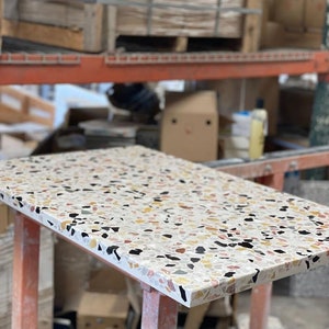 May include: A close-up of a white terrazzo table top with black, brown, and yellow flecks. The table top is on a set of orange metal legs.