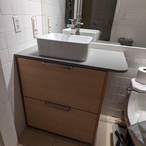 May include: A white bathroom vanity with two drawers and a white porcelain sink. The vanity is made of wood and has a light brown finish. The sink is rectangular and has a chrome faucet. The vanity is in a bathroom with white tiled walls and a beige tiled floor.