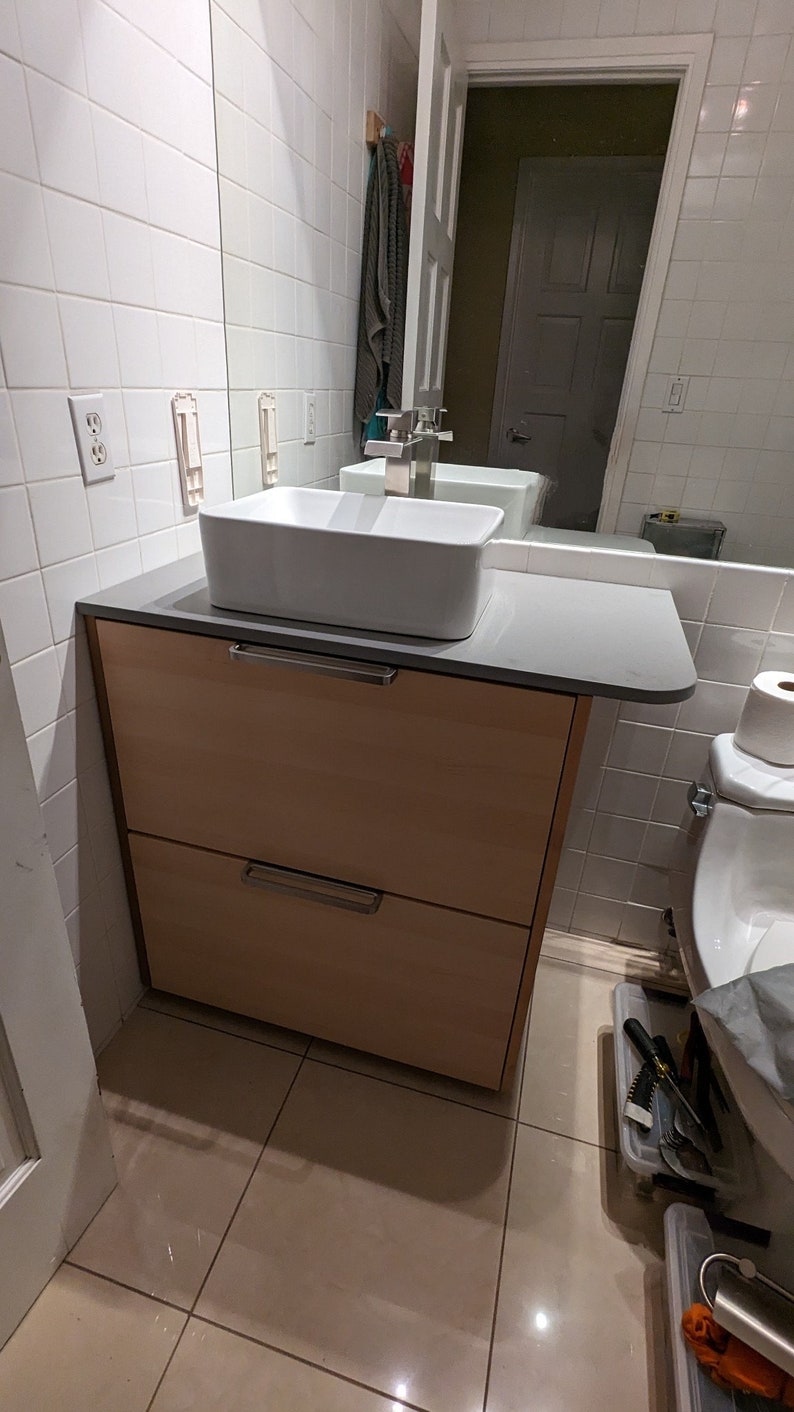 May include: A white bathroom vanity with two drawers and a white porcelain sink. The vanity is made of wood and has a light brown finish. The sink is rectangular and has a chrome faucet. The vanity is in a bathroom with white tiled walls and a beige tiled floor.
