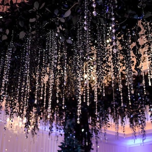 May include: An overhead floral arrangement with dark green leaves and hanging crystal strands. The crystals catch the light, creating a sparkling effect. The arrangement is likely used for event decoration, such as a wedding or party.