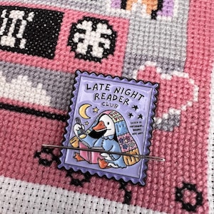 May include: A rectangular enamel needle minder featuring a purple postage stamp design with a duck reading a book under a blanket while holding a flashlight. The text reads LATE NIGHT READER CLUB and SLEEP IS OPTIONAL, BOOKS ARE NOT.