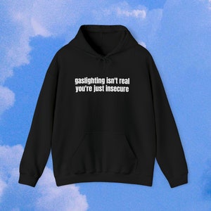 Op de afbeelding: Zwart hoodie met de tekst "gaslighting isn't real you're just insecure" in wit gedrukt op de voorkant.