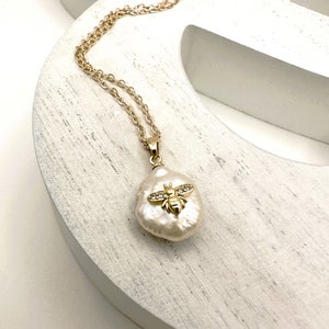 Puede incluir: Un collar de cadena dorada con un gran colgante de perla barroca blanca con una abeja dorada con detalles de cristal transparente.