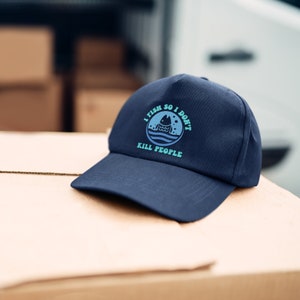 May include: Navy blue baseball cap with a curved brim. The cap features a circular graphic with a fish and waves, and the text "I FISH SO I DON'T KILL PEOPLE" in teal and white.