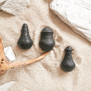 May include: Three small black ceramic figurines with faces and hats, sitting on a light brown sandy surface.
