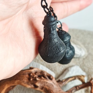 May include: Black earrings with a distinctive, bulbous form and textured finish, hanging from a black chain. The background features a hand, driftwood, and pale stones.