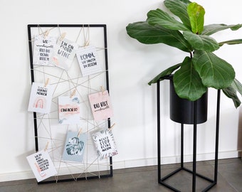 May include: A black metal grid wall display with various cards featuring motivational quotes and illustrations. The cards are held in place with clothespins and string. A potted plant with large green leaves sits on a black metal stand in the foreground.