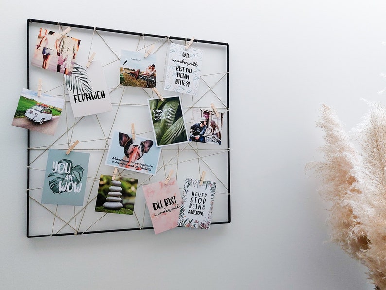 Op de afbeelding: Een zwart metalen rooster met touw en houten wasknijpers die verschillende ansichtkaarten met inspirerende citaten en afbeeldingen vasthouden. De citaten omvatten "You are wow", "Du bist wundervoll", "Never stop being awesome" en "Wie wundervoll bist du denn bitte?"