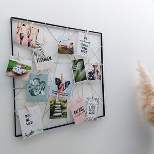 Op de afbeelding: Een zwart draadgaas met verschillende kaarten en foto's eraan bevestigd. De kaarten hebben inspirerende citaten en afbeeldingen van natuur, dieren en reizen. De foto's tonen een stel, een hond en een auto. Het gaas is bevestigd aan een witte muur.
