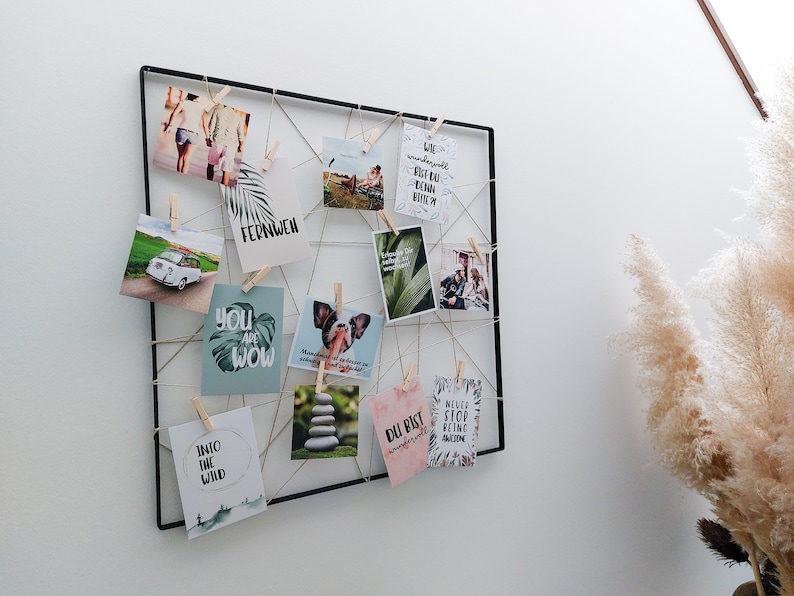 Op de afbeelding: Een zwart draadgaas met verschillende kaarten en foto's eraan bevestigd. De kaarten hebben inspirerende citaten en afbeeldingen van natuur, dieren en reizen. De foto's tonen een stel, een hond en een auto. Het gaas is bevestigd aan een witte muur.