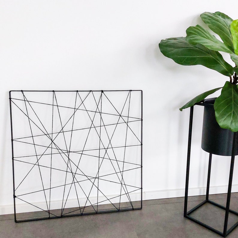 Op de afbeelding: Een zwarte metalen wanddecoratie met een geometrisch ontwerp van elkaar kruisende lijnen. De decoratie hangt aan een witte muur.