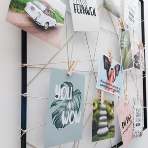 Op de afbeelding: Een zwart geframed prikbord met touw en wasknijpers die verschillende kaarten met inspirerende citaten en afbeeldingen vasthouden. De citaten omvatten "You are Wow", "Du bist wundervoll", "Never stop being awesome", "Fernweh" en "Manchmal ist es besser zu schweigen und zu l&auml;cheln".