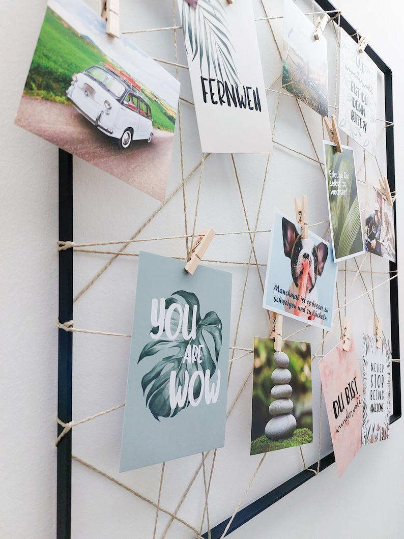 Op de afbeelding: Een zwart geframed prikbord met touw en wasknijpers die verschillende kaarten met inspirerende citaten en afbeeldingen vasthouden. De citaten omvatten "You are Wow", "Du bist wundervoll", "Never stop being awesome", "Fernweh" en "Manchmal ist es besser zu schweigen und zu l&auml;cheln".