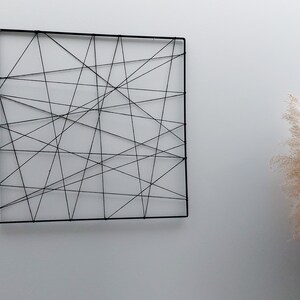 Op de afbeelding: Een zwarte vierkante wanddecoratie gemaakt van touw, met een geometrisch patroon van elkaar kruisende lijnen.