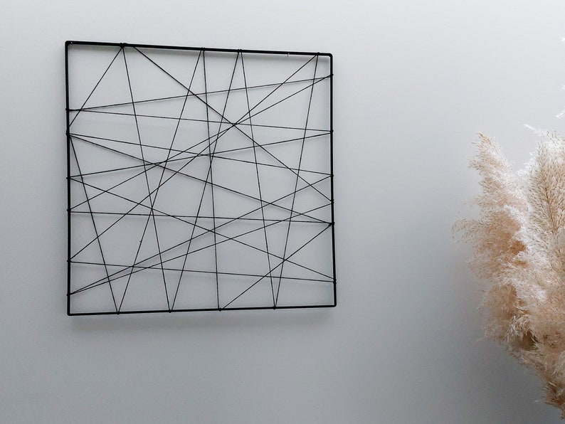 Op de afbeelding: Een zwarte vierkante wanddecoratie gemaakt van touw, met een geometrisch patroon van elkaar kruisende lijnen.