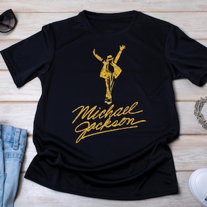 May include: Black t-shirt featuring a gold glitter design of Michael Jackson in a signature pose, with his name in a matching gold script. The shirt is displayed with sunglasses, jeans, and sneakers.