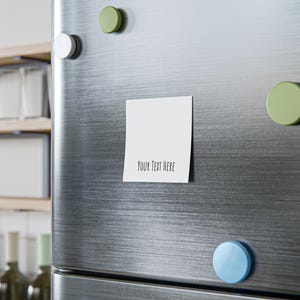 May include: A white square sticky note with the text "YOUR TEXT HERE" in a black, handwritten-style font. The note is attached to a stainless steel refrigerator door with colorful round magnets.