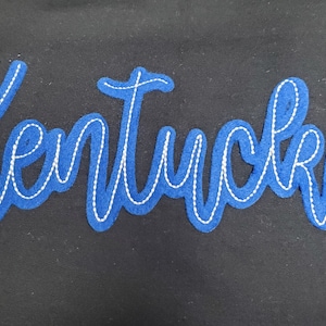 May include: A blue felt applique with the word "Kentucky" in a cursive script, outlined with white stitching, is centered on a black background. The design is simple and bold, suitable for apparel or crafts.