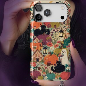 Pode incluir: Uma capa para iPhone 17 Pro Max com acabamento brilhante, apresentando uma ilustração colorida com tema de outono, contendo gatos pretos, um pequeno fantasma, abóboras laranjas e verdes, casas, lanternas, cogumelos e plantas em fundo creme.