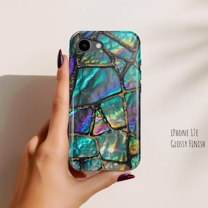 以下が含まれることがあります： 光沢仕上げのiPhone 17E用スマホケース。ティール、ブルー、パープルの色合いの虹色のアバロンシェルの破片がモザイク状に配置され、暗く細い不規則な線で区切られた石のようなデザインが特徴です。