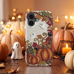 Op de afbeelding: Een telefoonhoesje met een geborduurd ontwerp op een crèmekleurige stoffen achtergrond. Het patroon bevat oranje pompoenen, groene en bruine herfstbladeren, rode bessen en twee kleine witte spookjes met eenvoudige zwarte ogen en monden.