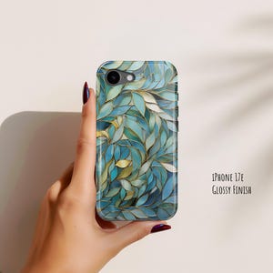 Puede incluir: Una funda para iPhone 17E con acabado brillante, que presenta un patrón denso de hojas superpuestas en tonos azul verdoso, azul y crema, delineadas con finas líneas doradas que cubren toda la superficie posterior del dispositivo.