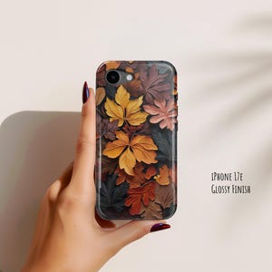 May include: A phone case for an iPhone 17E with a glossy finish, featuring a dense, overlapping pattern of autumn leaves in shades of yellow, orange, deep red, brown, and dark gray. The design covers the entire back of the rectangular device case.