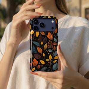 May include: A smartphone case featuring a repeating pattern of autumn leaves in shades of orange, yellow, brown, and muted blue against a black background. The case has a dark blue camera module cutout with three circular lenses and a flash.
