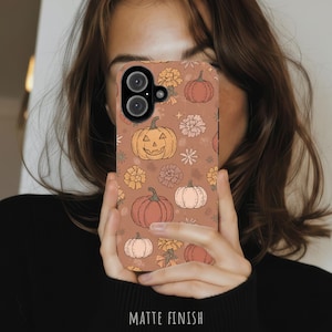 May include: A phone case with a matte finish featuring a repeating pattern of orange and cream-colored pumpkins and various flowers on a terracotta-colored background. The design includes a jack-o'-lantern with a carved face and small stars.
