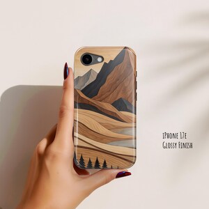 May include: A phone case with a glossy finish featuring a stylized landscape illustration of brown and tan mountains, rolling hills, and black pine trees at the bottom. The text on the right reads iPhone 17E Glossy Finish.