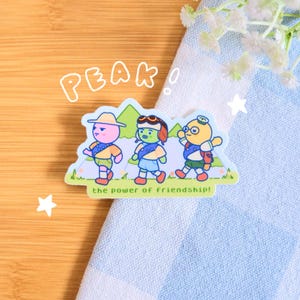 以下が含まれることがあります： スカウトの制服を着た3人の漫画のキャラクターが一緒に歩いているステッカー。ステッカーには、バブルフォントで「PEAK」という単語と、下部に「the power of friendship！」というフレーズがあります。キャラクターはピンク、緑、黄色です。