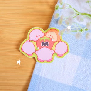 以下が含まれることがあります： 漫画のキャラクターのグループが描かれたダイカットステッカー。中央のキャラクターは、トマトの形をした頭を持つピンク色のフィギュアです。その周りには、笑顔の顔をした他のピンクとオレンジのキャラクターがいます。ステッカーには緑色の縁取りがあります。