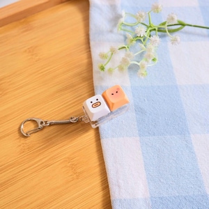 May include: A novelty keychain featuring two keyboard keycaps with cartoon faces. One keycap is white with a surprised face, and the other is peach-colored with a smiling face. The keychain has a silver-colored spring and clasp.