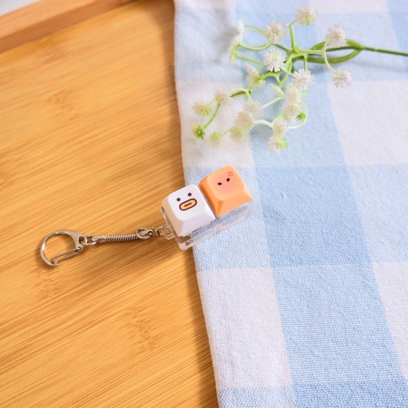 May include: A novelty keychain featuring two keyboard keycaps with cartoon faces. One keycap is white with a surprised face, and the other is peach-colored with a smiling face. The keychain has a silver-colored spring and clasp.