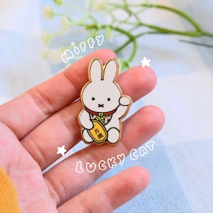 Op de afbeelding: Een wit konijn met een rode halsband en een geel bord met de tekst "招財" houdt een gouden munt vast. Het konijn is een gelukkatten-hanger en maakt deel uit van een Miffy-collectie.