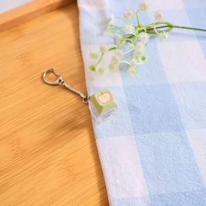 May include: A keychain featuring a green square charm with a red apple design. The charm is attached to a silver-colored chain and clasp. The background includes a blue and white checkered cloth and a wooden surface.
