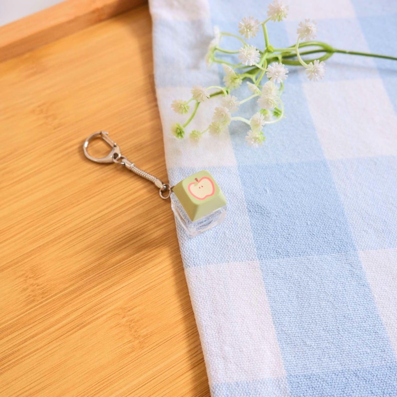May include: A keychain featuring a green square charm with a red apple design. The charm is attached to a silver-colored chain and clasp. The background includes a blue and white checkered cloth and a wooden surface.