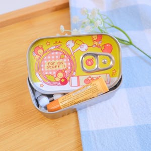 May include: A yellow sardine-shaped tin with the text "for my stuff :)" and illustrations of cherubs, earphones, and a lip balm. The tin is open, revealing a lip balm and a pair of white earphones.