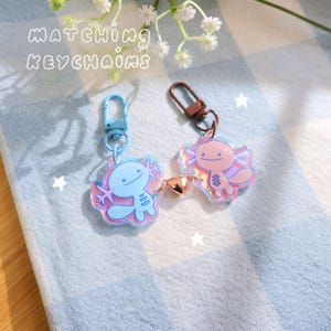 May include: Two axolotl keychains. One is light blue, the other is pink, both with iridescent accents. Each keychain has a metal clasp and a small heart charm. The text "MATCHING KEYCHAINS" is above the keychains.
