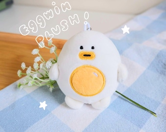 Puede incluir: Un peluche blanco con forma de huevo redondo con una barriga circular amarilla, ojos pequeños negros y un pico amarillo. El texto "eggwin plush" está escrito en letras de burbuja blancas sobre el juguete, junto a dos pequeñas estrellas blancas.