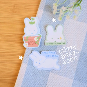 Może przedstawiać: Trzy białe naklejki z królikami z różnymi napisami. Pierwsza naklejka przedstawia królika w doniczce z napisem "Wciąż rosnę". Druga naklejka przedstawia królika z napisem "Koniec pracy". Trzecia naklejka przedstawia leżącego królika z napisem "Leniwe dni są w porządku". Naklejki znajdują się na niebiesko-białym wzorze w kratkę z napisem "Miffy Self-Care" w kolorze białym.