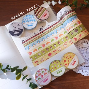 Puede incluir: Una colección de rollos de cinta decorativa washi con varios patrones, incluyendo dinosaurios pixelados, pingüinos azules, fresas, setas y frutas cortadas. Los rollos se muestran sobre un cuaderno cuadriculado con tiras de cinta adhesiva.