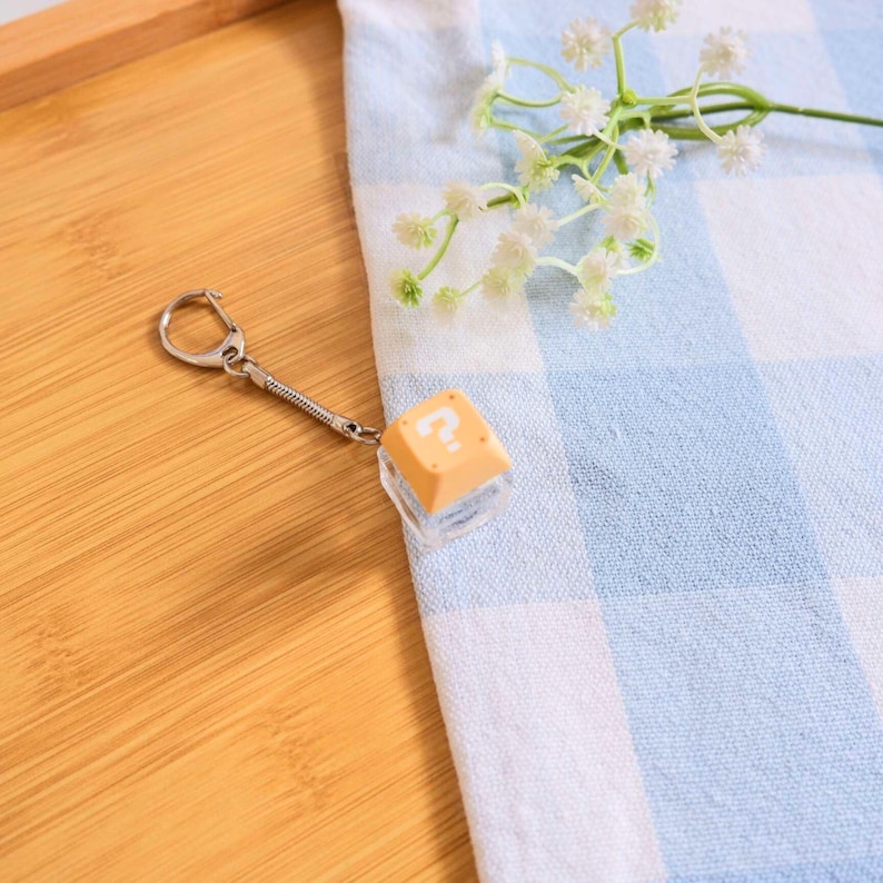 May include: A keychain with a clear, cube-shaped charm topped with a light orange button featuring a white question mark. The keychain has a silver-colored clasp and a coiled metal chain. The background includes a blue and white checkered cloth and a wooden surface.