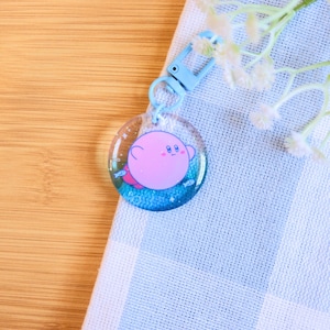 May include: A pink and blue keychain featuring a cartoon character with a round body and large eyes. The character is surrounded by blue and white glitter and small fish.