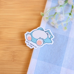 May include: A blue and pink sticker with a cartoon of a sleeping Pokemon character. The sticker says "You deserve a nap".