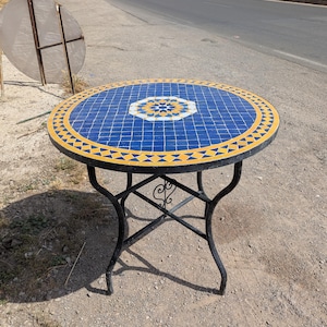 May include: Round outdoor table with a mosaic tile top. The tabletop features a blue and white geometric design with a yellow border. The table has a black metal base with curved legs.