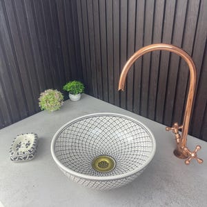 May include: A white ceramic vessel sink with a black geometric pattern, a copper faucet, and a matching soap dish. The sink has a gold-colored drain and is set on a gray surface. Small potted plants are in the background.